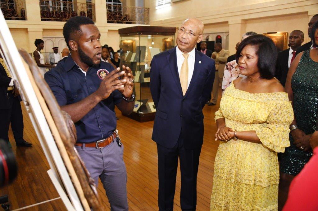Their Excellencies Sir Patrick Allen and Lady Allen listen attentively as Mr. Ramsay of the Institute of Jamaica expounds on the significance of the sculpture, “From Rude Boy to Rastafari: From Rocksteady to Reggae”, made by Rass Ruggles (Winston Green) during the tour of an exhibition at King’s House on the occasion of The Governor-General’s Reception marking Jamaica 55 Independence Celebration.
